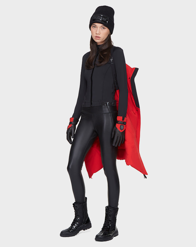 A woman poses against a light backdrop, draping the Rudsak ELISA - L121512 POPPY RED sustainable winter jacket over her shoulder, styled with a black beanie, long-sleeve top, faux leather leggings, gloves, and boots for a sleek look.