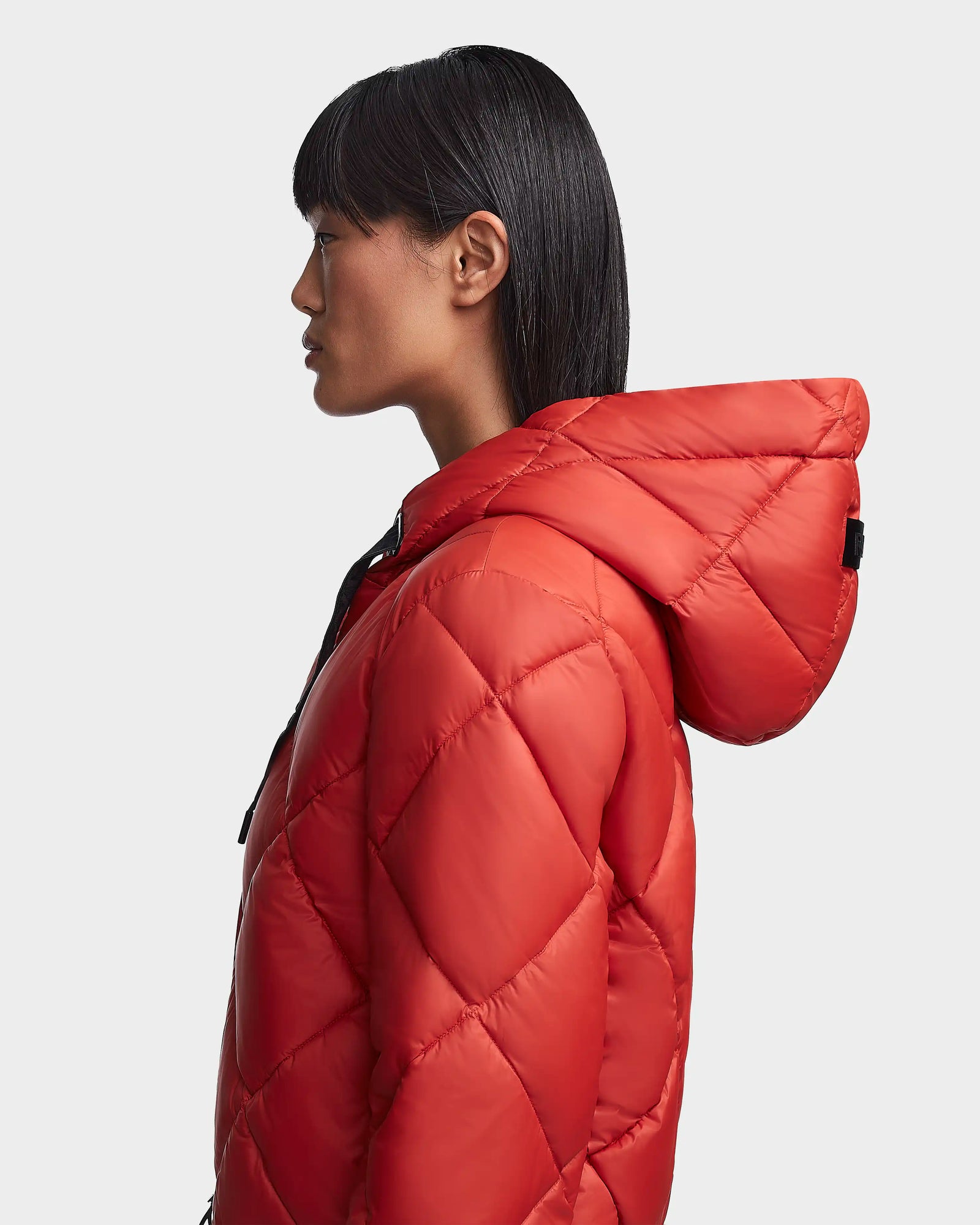A woman with straight, dark hair and bangs is shown in profile wearing the Rudsak BALA - 8123614 Orange sustainable puffer jacket against a plain light gray background.