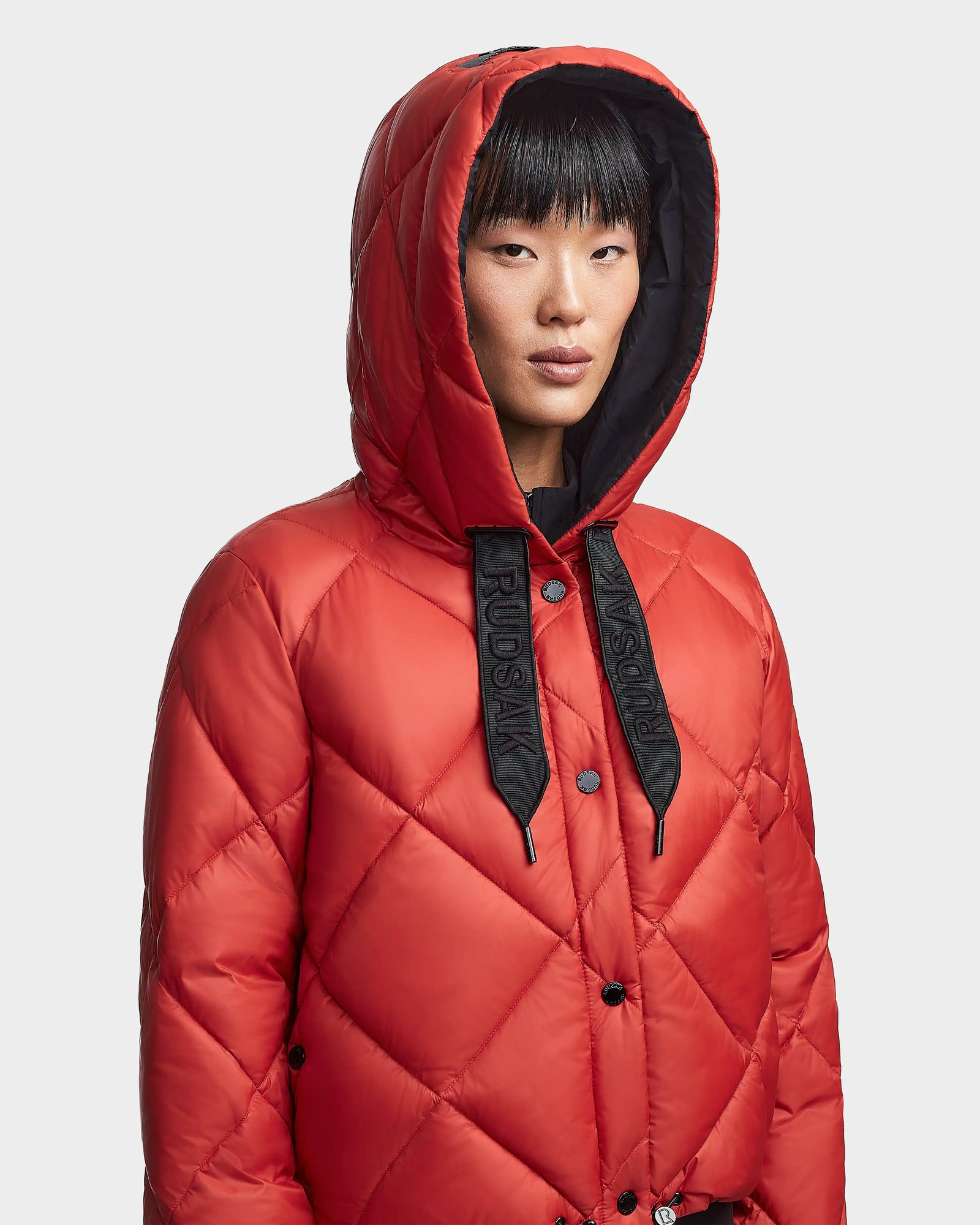 A person wears the Rudsak BALA - 8123614 Orange women's quilted winter coat with black "Rudsak" drawstrings, standing against a plain light background.