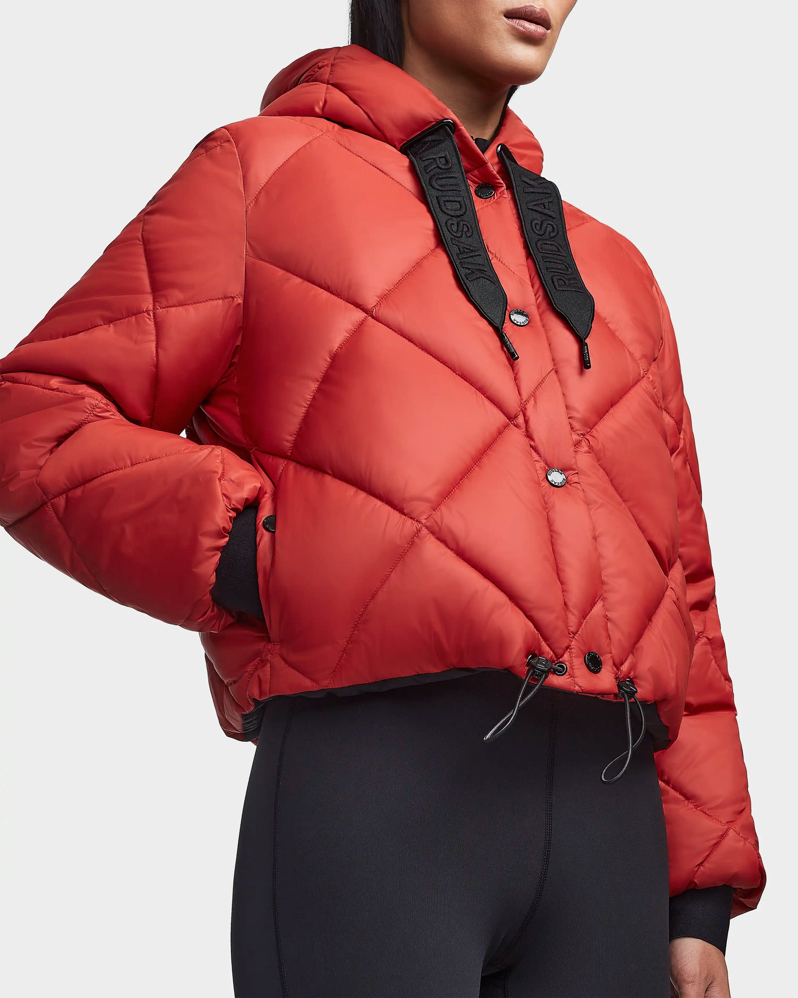 A woman wears the Rudsak BALA - 8123614 Orange sustainable puffer jacket with a hood, black drawstrings, snap buttons, and black ribbed cuffs, paired with black leggings.