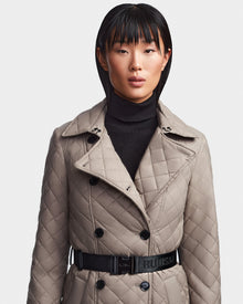 A person with straight, dark hair and blunt bangs wears the Rudsak DAISY - 8123580 Cement women's quilted maxi trench coat with a belted waist over a black turtleneck, standing against a plain light background.