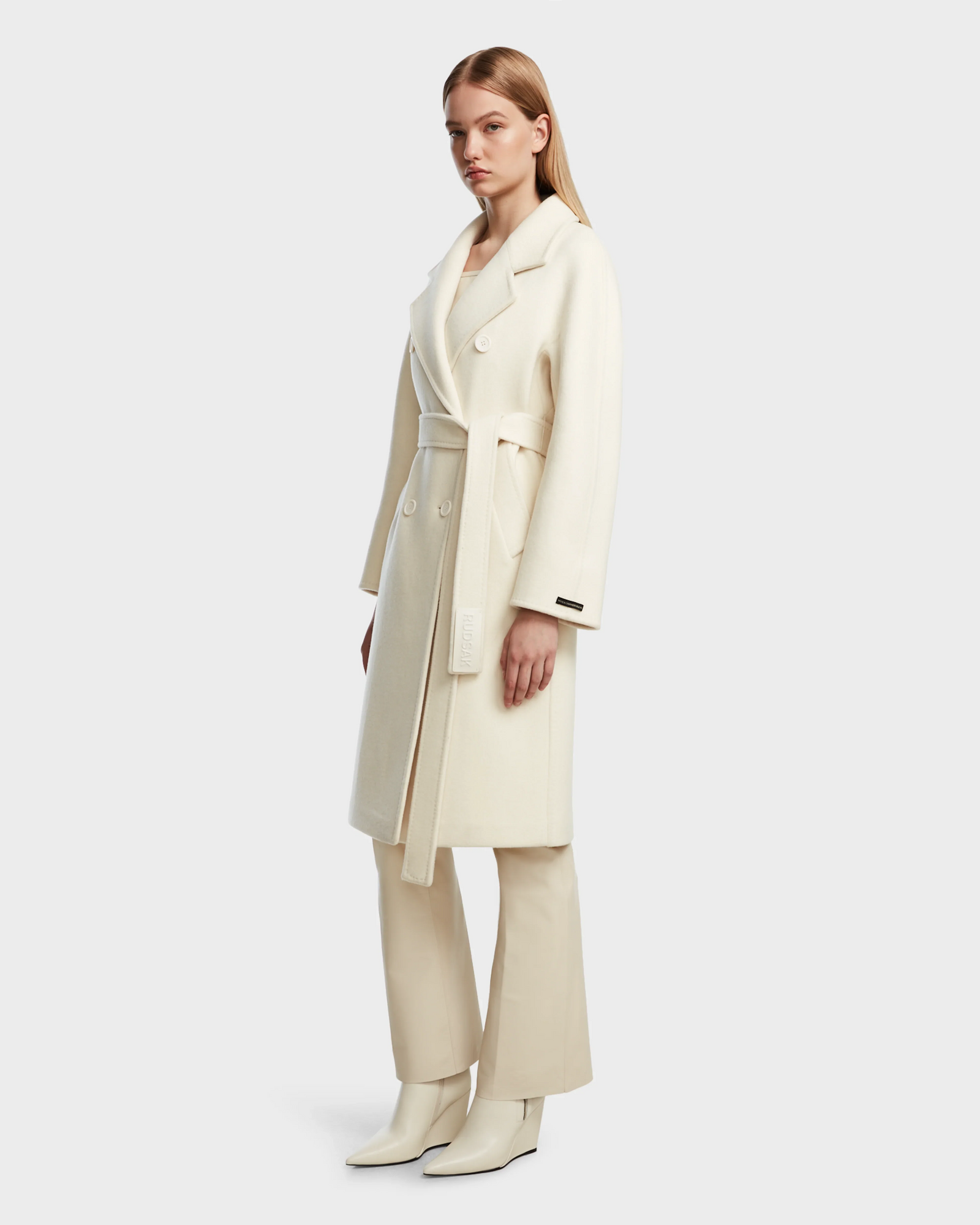 A woman wears the Rudsak KOURT W - 8124702 Iceland cream belted wool coat with wide lapels, paired with cream pants and white ankle boots, standing against a plain light background.