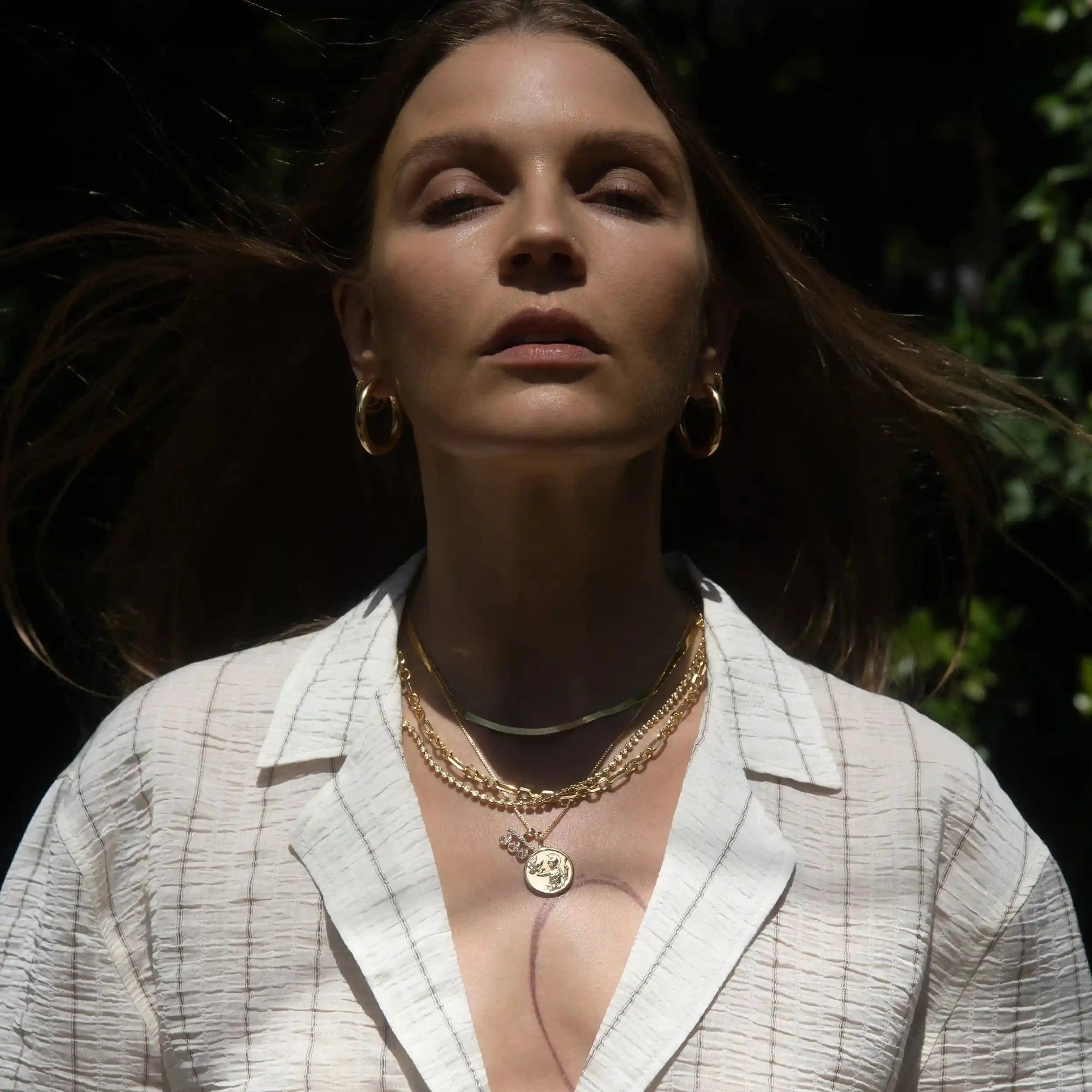 A person with long brown hair wears gold hoop earrings and the Awe Inspired Statement Chain Necklace, standing outdoors in sunlight, with eyes half-closed, wearing a white textured top.