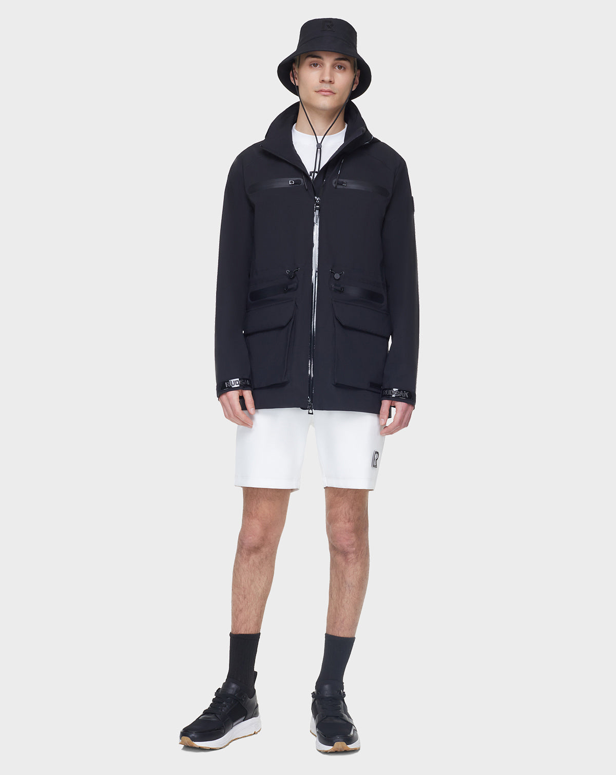 A young man stands facing forward, wearing the RUDSAK JACY - 6122000 BLACK mens rain trench coat over a black bucket hat, jacket, white shorts, black crew socks, and sneakers against a plain light background.
