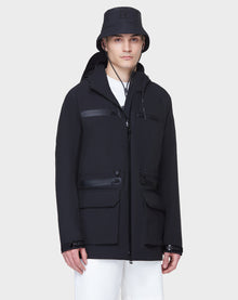 A person in the RUDSAK JACY - 6122000 BLACK water-resistant trench with multiple zippered pockets and a matching black bucket hat stands against a light gray background, wearing white pants and a neutral expression.