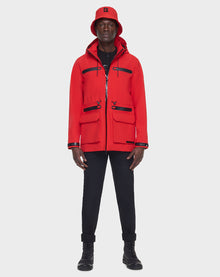 A person faces forward wearing the RUDSAK JACY - 6122000 POPPY RED mid-length raincoat with black zippers and pockets, a matching red bucket hat, black pants, and black boots against a plain light background.