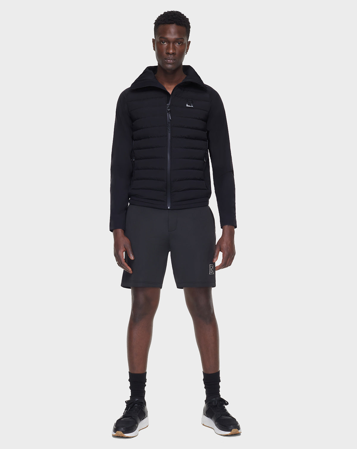 A man faces forward against a white background, wearing the RUDSAK JARRY - 6122007 BLACK men’s windbreaker jacket with black shorts, socks, and sneakers—a versatile cold-weather essential.
