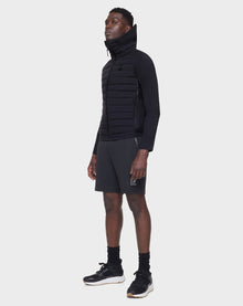 A man stands against a plain background wearing the RUDSAK JARRY - 6122007 BLACK high-collar puffer vest over a black long-sleeve top, paired with dark athletic shorts, black socks, and sneakers. He faces slightly sideways with a neutral expression.