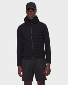 A person wears the RUDSAK JARRY - 6122007 BLACK men’s padded windbreaker jacket with a hood, paired with black shorts and a baseball cap, standing against a plain white background—a cold-weather staple.