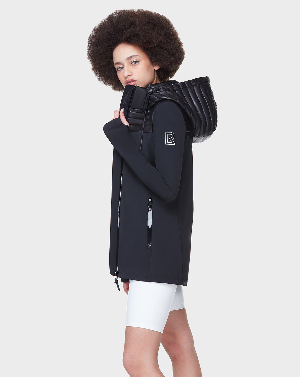 A person with a curly afro wears the Rudsak CAMIRA - 8122011 BLACK activewear jacket, featuring waterproof zippers and a padded collar, paired with white shorts, against a plain light gray background.